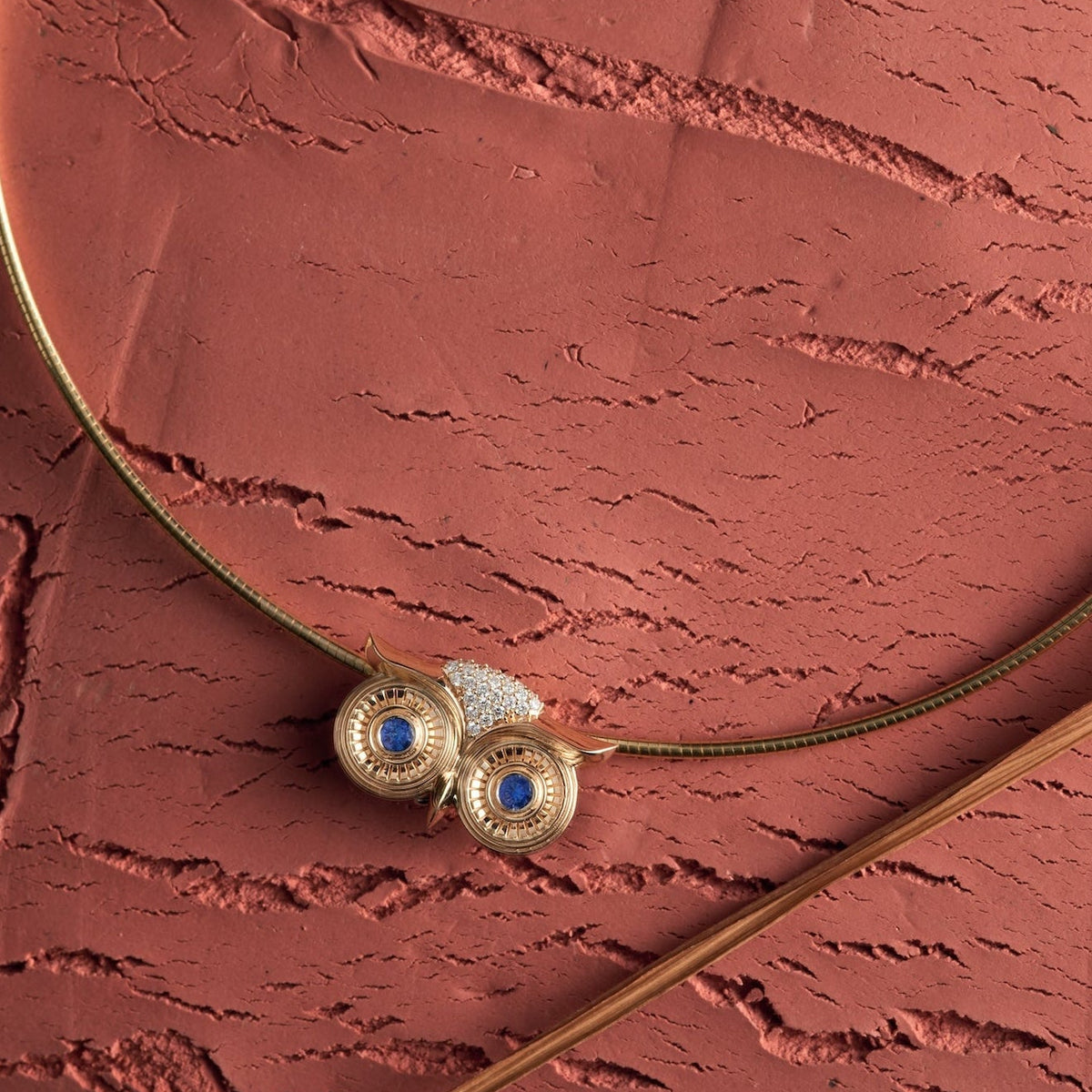 The SELIN KENT Athena gold owl necklace with blue sapphire eyes laid flat in a circular omega chain on a textured terracotta surface